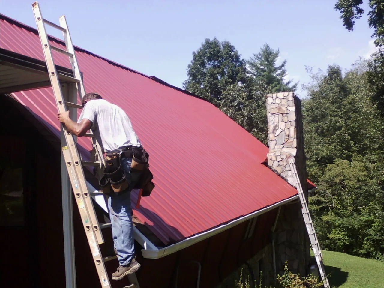 Roofer on a ladder beside a red metal roof and stone chimney — Soffit & Fascia Repair by Roofing Brothers in Fielding